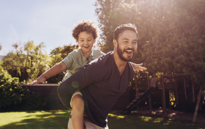 Un padre llevando a su hijo a cuestas en un día cálido de verano, ambos sonriendo y disfrutando del momento.