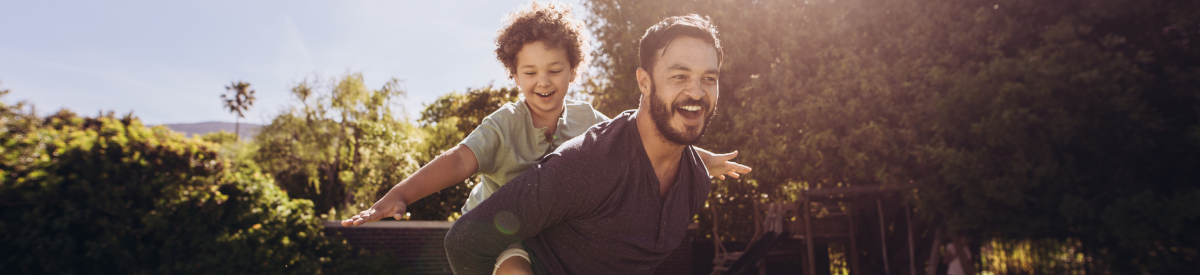 Un padre llevando a su hijo a cuestas en un día cálido de verano, ambos sonriendo y disfrutando del momento.